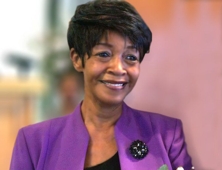 Smiling woman in a purple blazer with a black brooch against a blurred indoor background