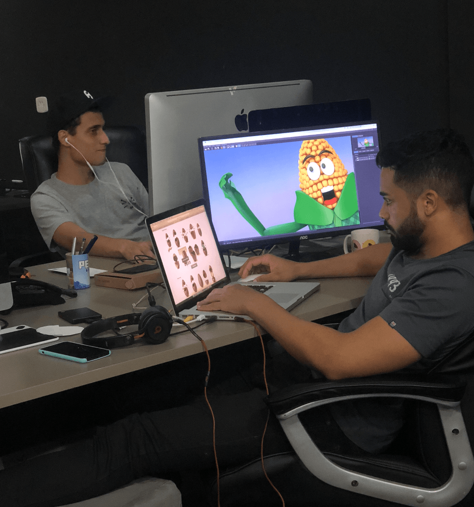 Dois homens trabalham em uma mesa com computadores. Um olha para uma tela com um personagem de desenho animado de milho, o outro olha para a tela de um laptop.