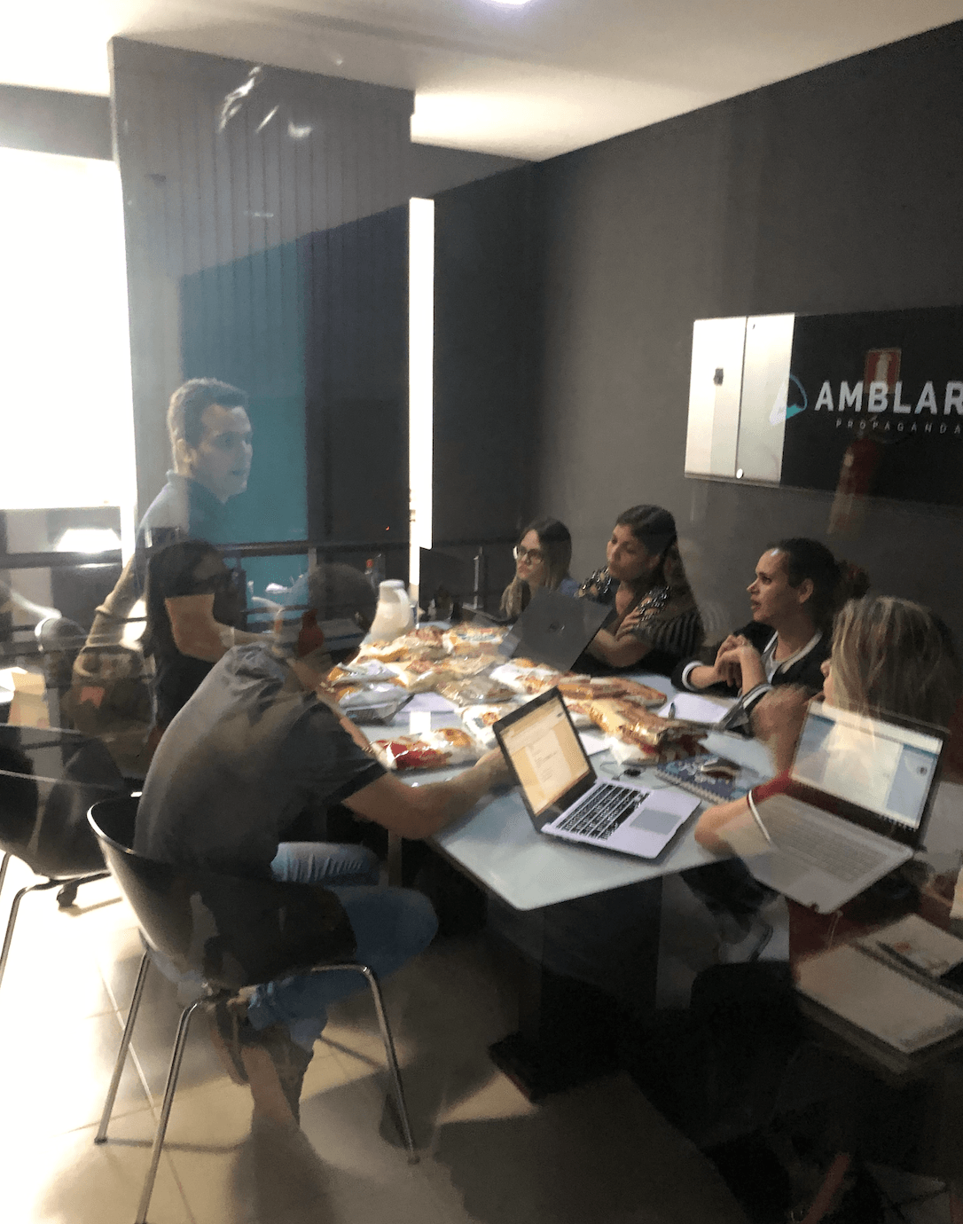Pessoas em uma sala de reuniões, trabalhando em laptops e papéis ao redor de uma mesa. Uma pessoa em pé fala. Logotipo