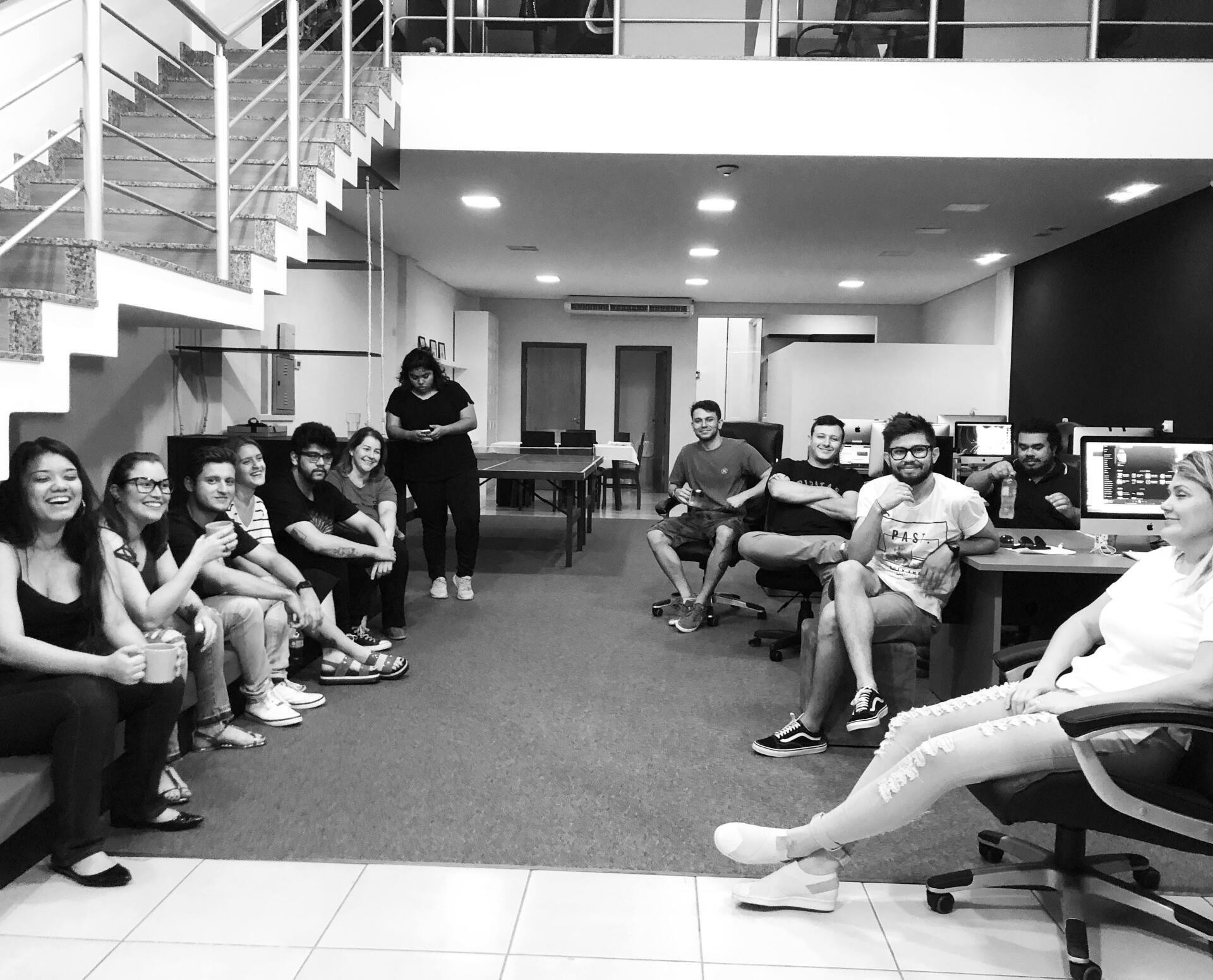 Um grupo de pessoas sentadas e em pé em um escritório. Algumas sorriem, e a cena é em tons de cinza.