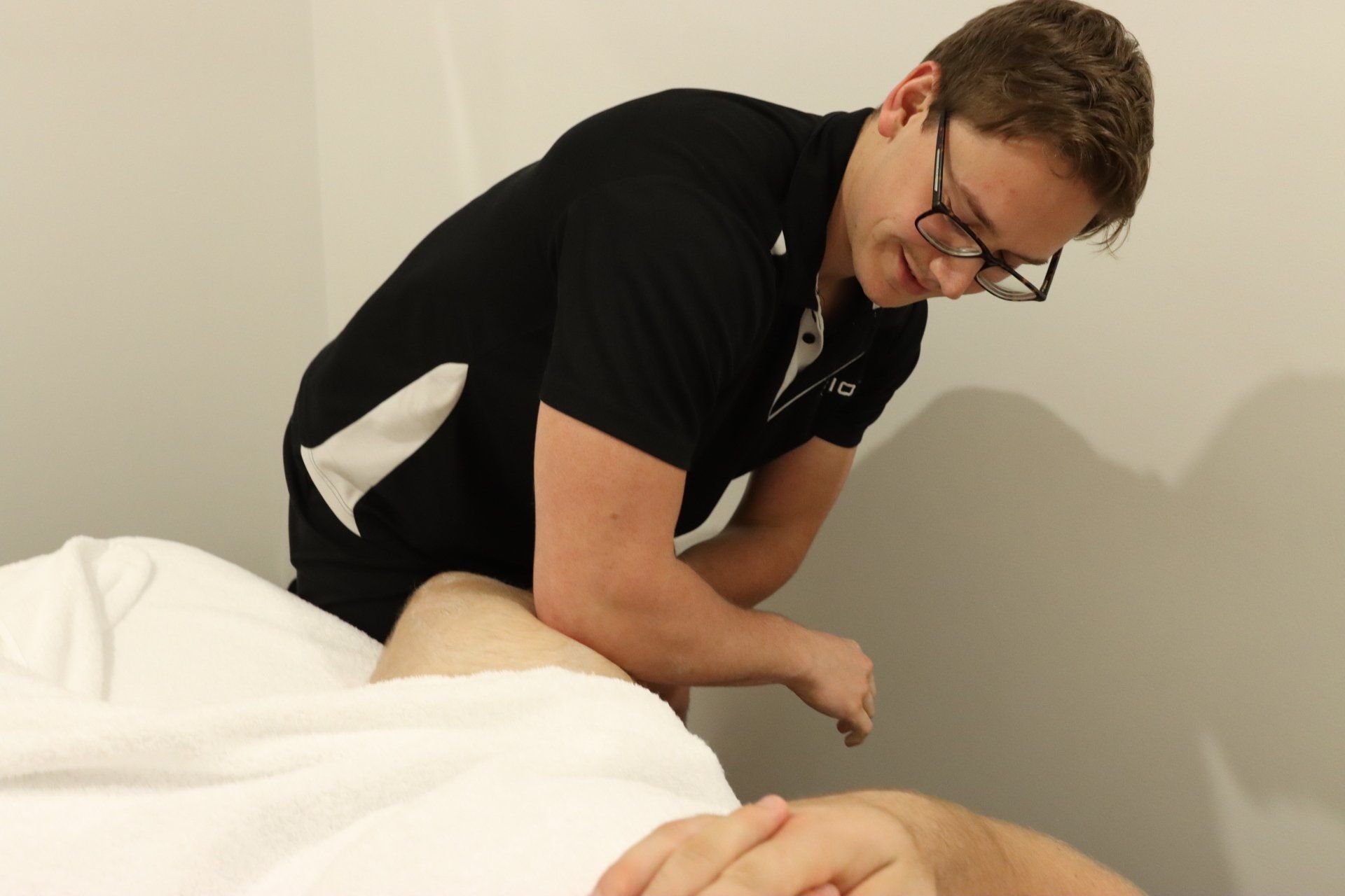 Man giving leg massage in a neutral-toned room. He is wearing glasses and a black polo.