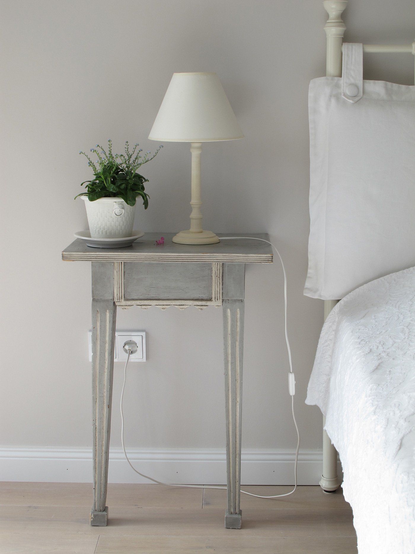 A small, weathered gray wooden bedside table with a lamp and a potted plant sits next to a white bed against a gray wall.