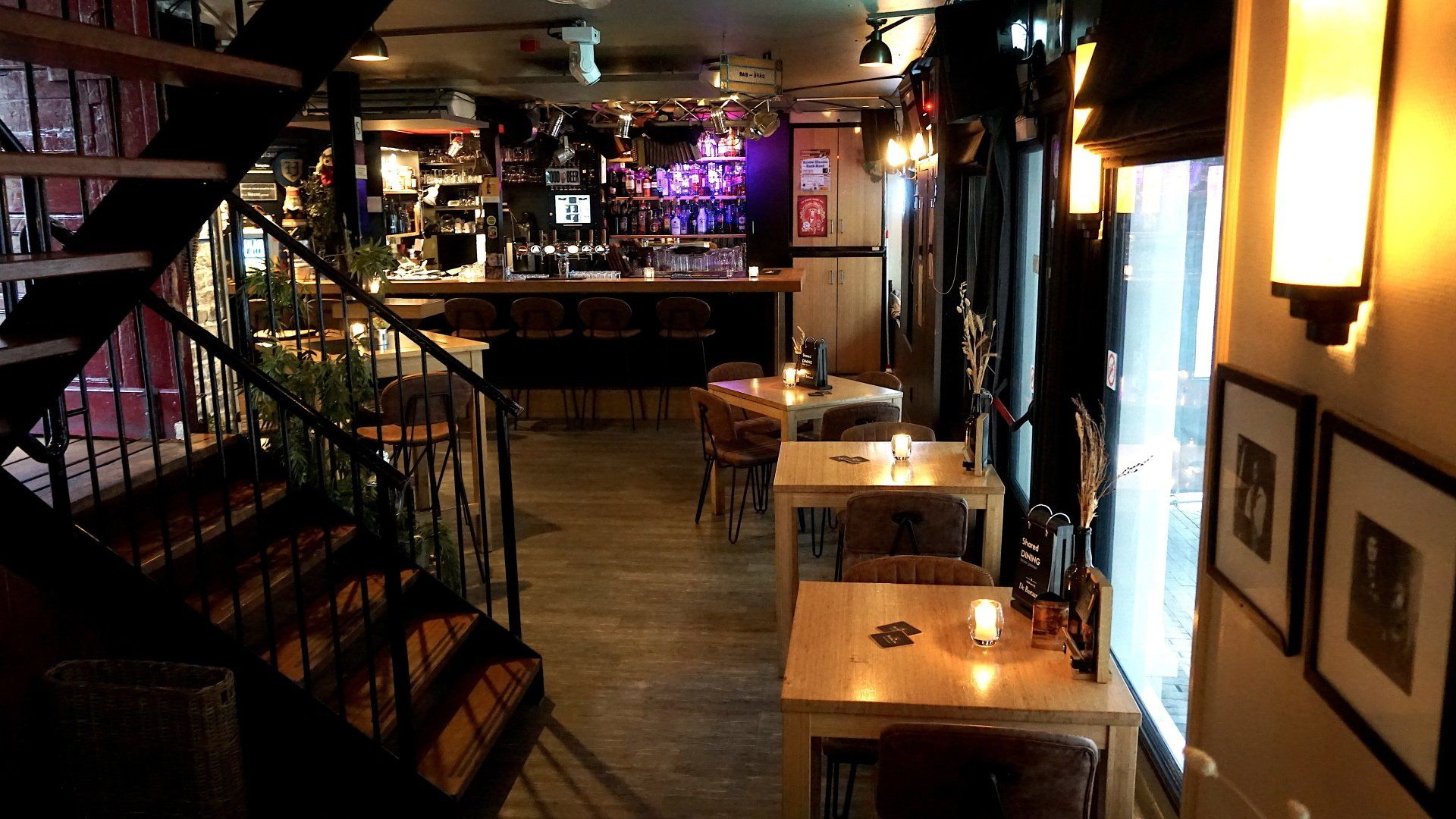 Gezellige bar met tafels, barkrukken, een trap, warme verlichting en een podium achterin.