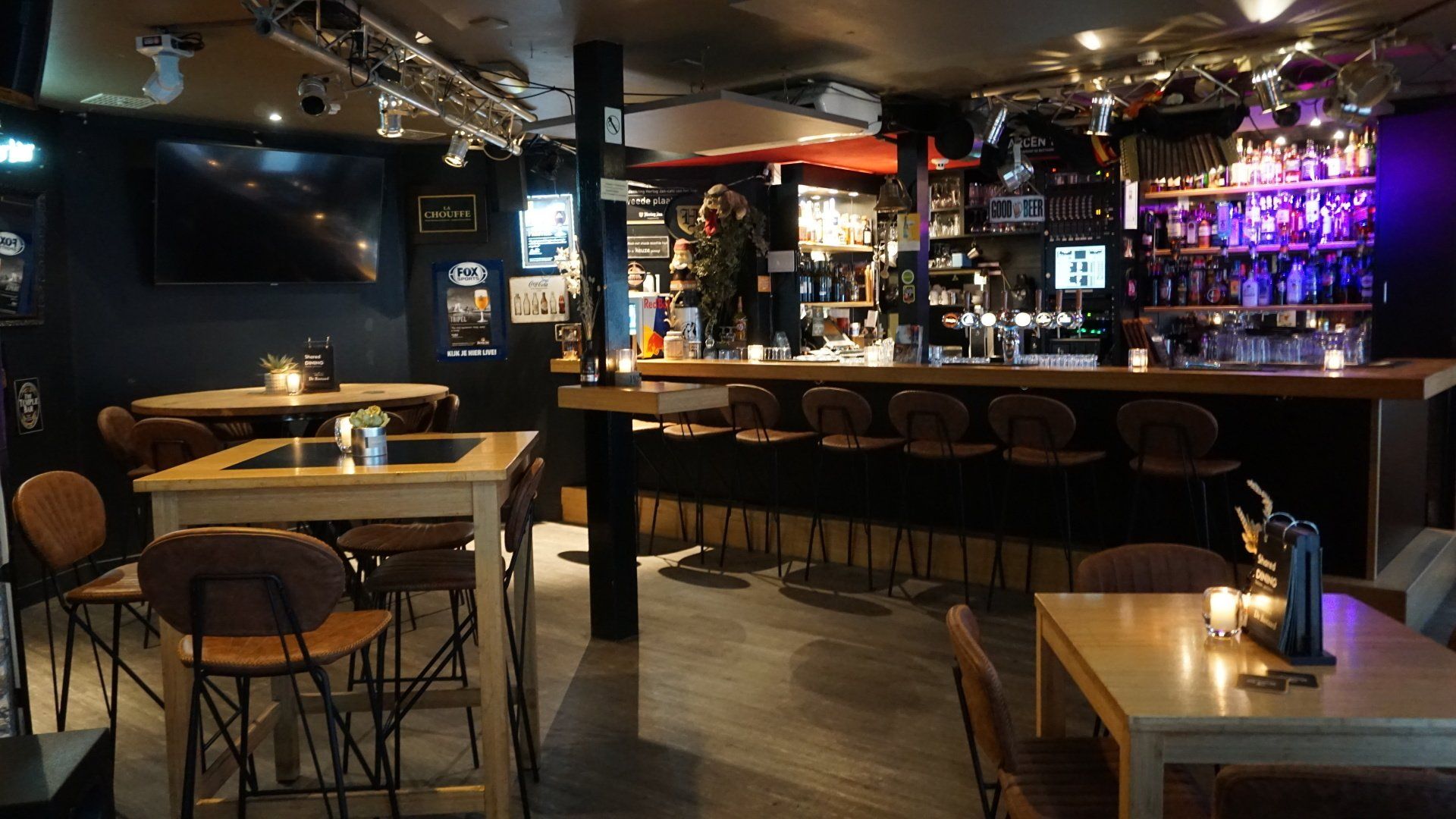 Een schemerig verlichte bar met houten tafels, barkrukken en een lange toonbank vol flessen en televisies.