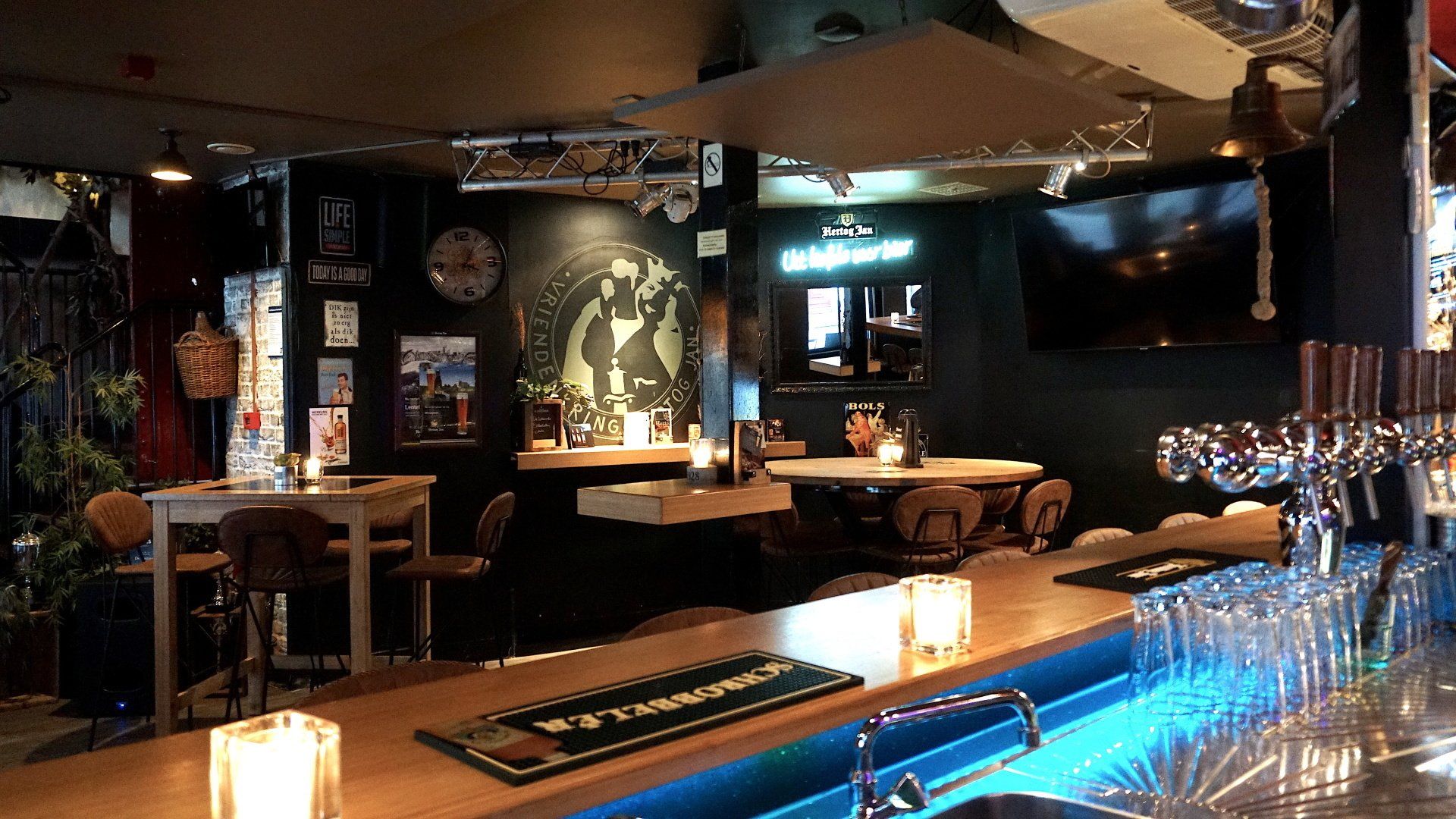 Een sfeervol verlichte bar met houten tafels, barkrukken, een bar en warme, decoratieve verlichting.