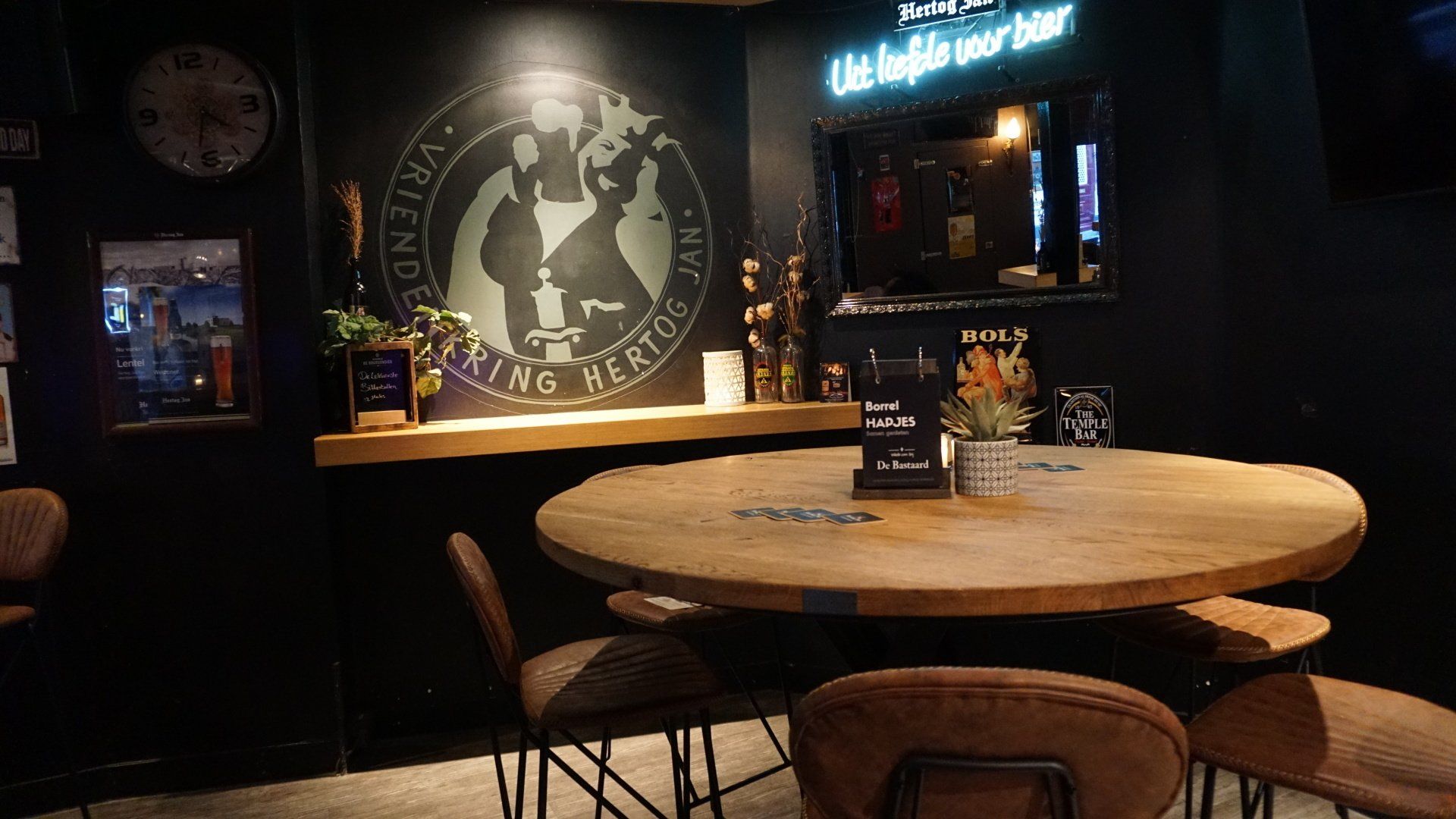 Een schemerig barinterieur met ronde houten tafels, donkere muren, muurdecoratie en neonreclame.