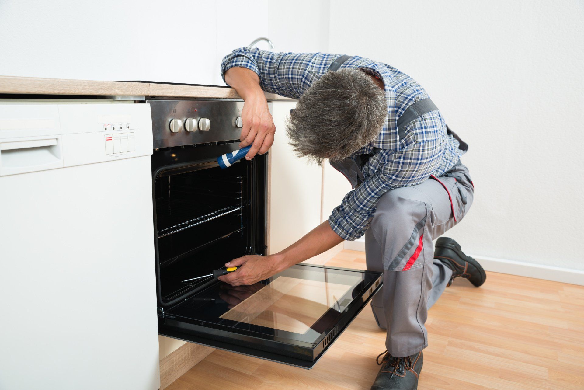 A man is fixing an oven in a kitchen with a screwdriver.