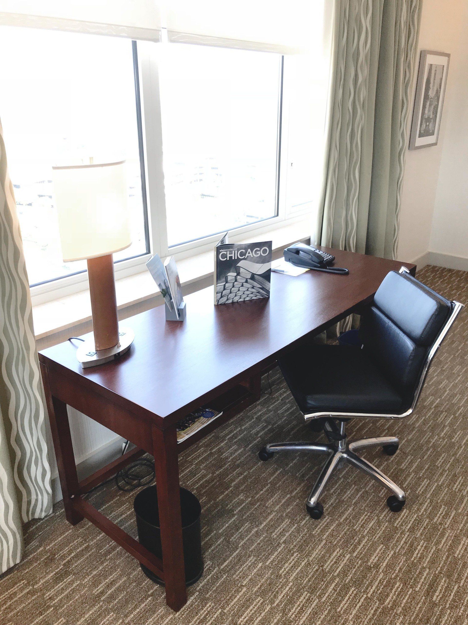 A wooden desk with a black chair in front of a window.