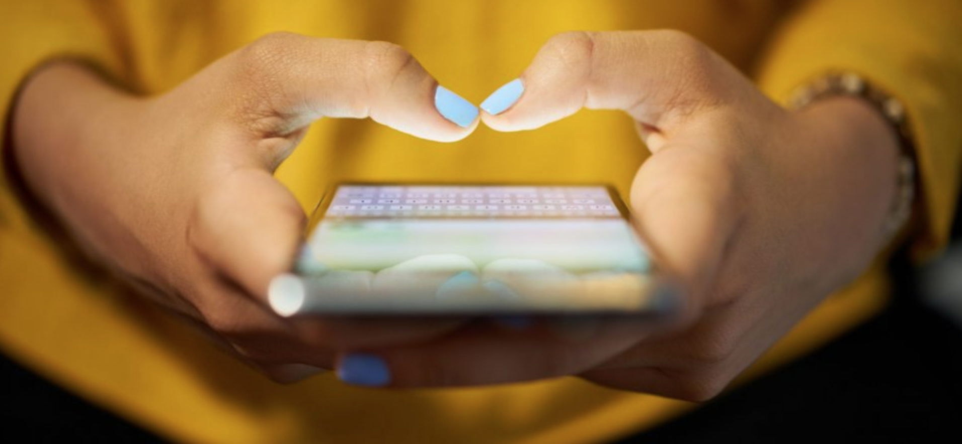 A woman is holding a cell phone in her hands and making a heart shape with her fingers.