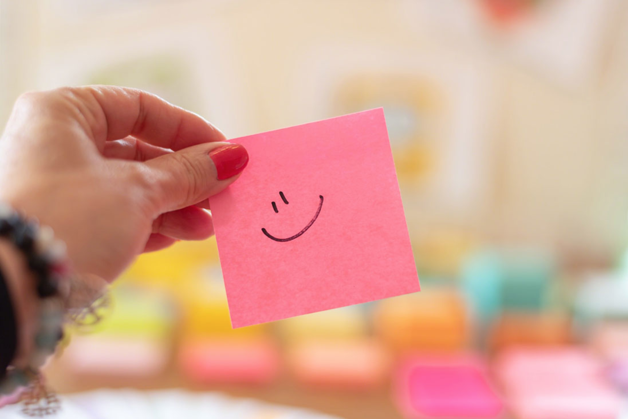 A person is holding a pink sticky note with a smiley face drawn on it.
