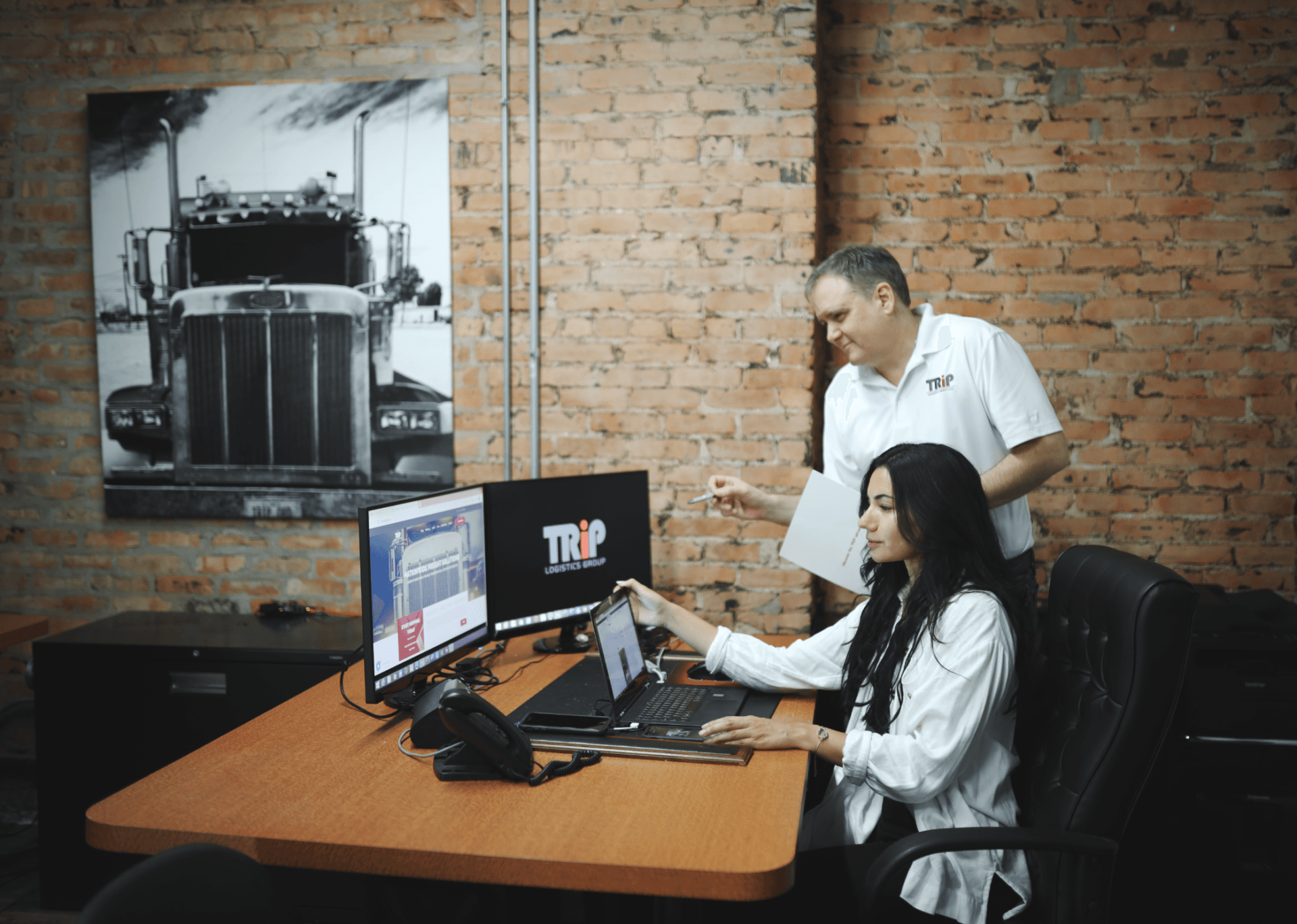A man and a woman are sitting at a desk looking at a laptop.