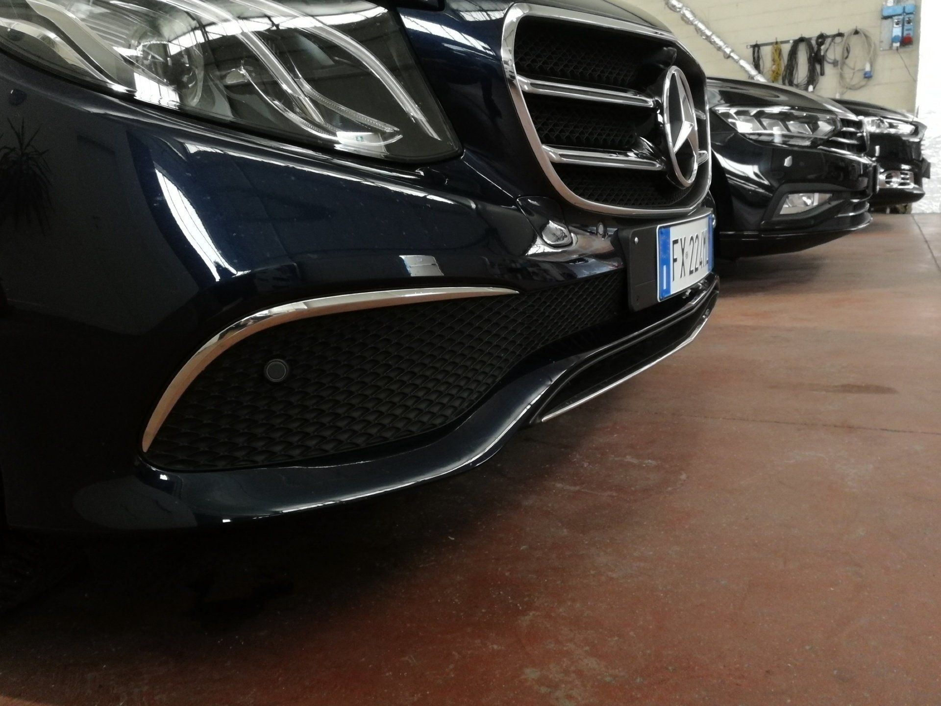 Primo piano del paraurti anteriore di una Mercedes-Benz blu, parcheggiata in un garage. Sullo sfondo si vedono altre auto.