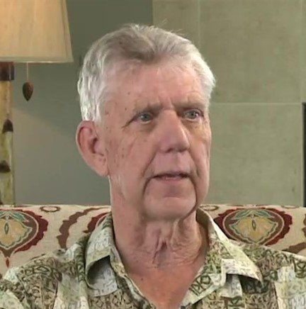 An older man is sitting on a couch with a lamp in the background.
