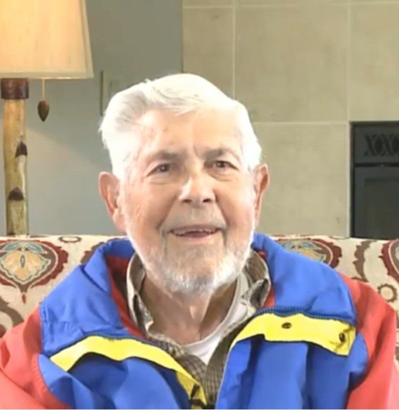An older man wearing a blue and yellow jacket sits on a couch