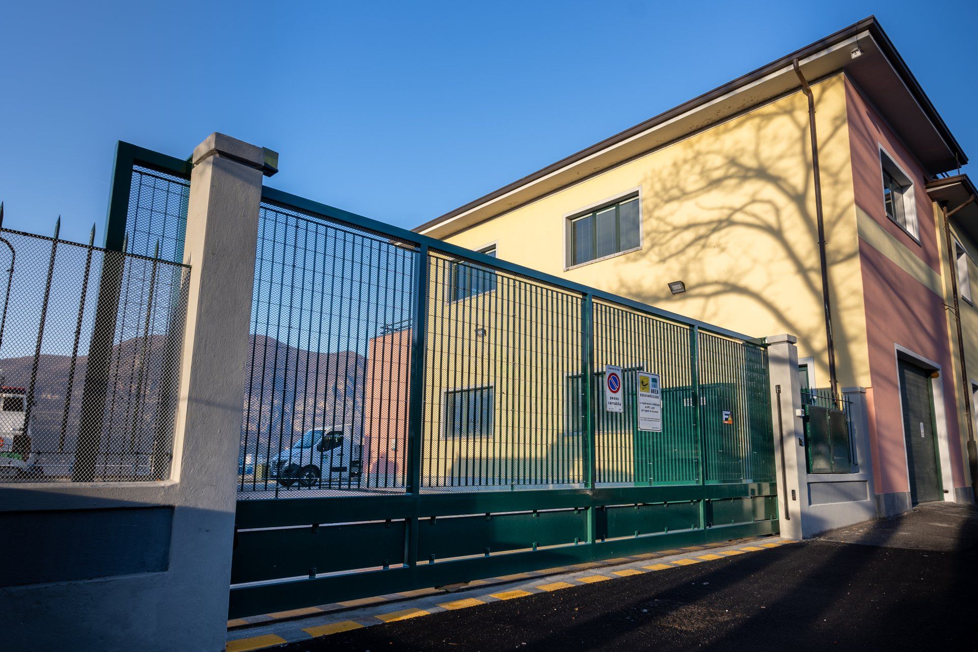 Un cancello di metallo verde accanto a un edificio giallo sotto un cielo azzurro, con le ombre degli alberi spogli sul muro.