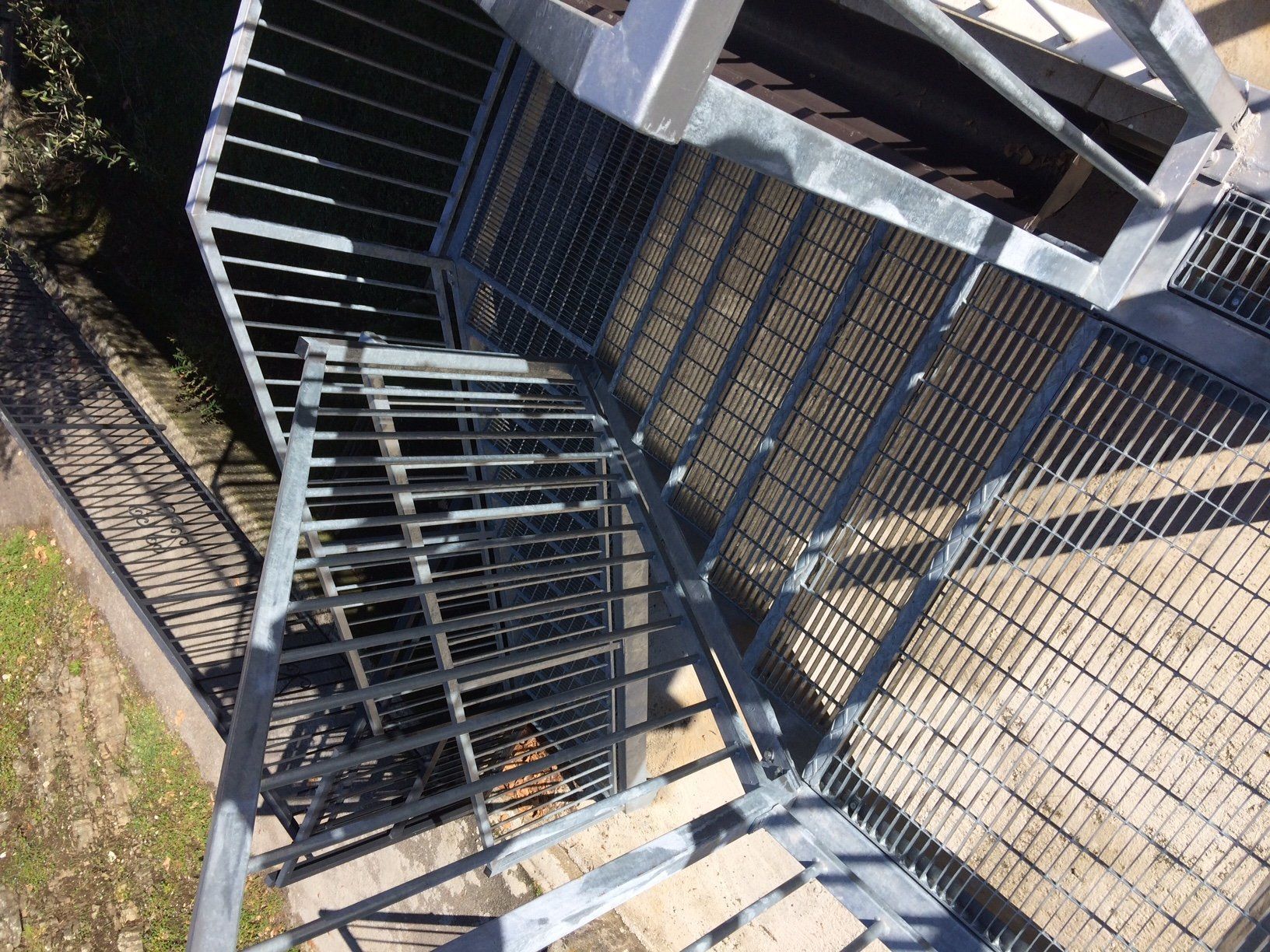 Scala in metallo e ringhiera nera accanto a una recinzione a rete metallica su un soleggiato vialetto esterno.