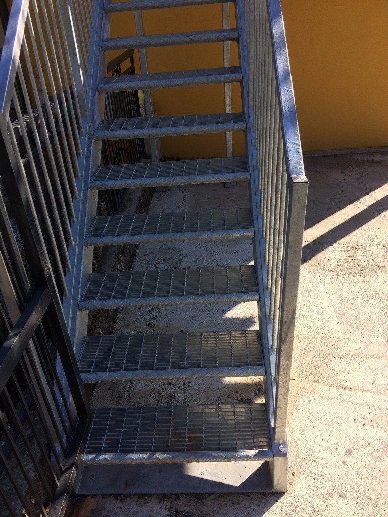 Scala metallica con gradini grigliati e ringhiere nere accanto a un edificio giallo.