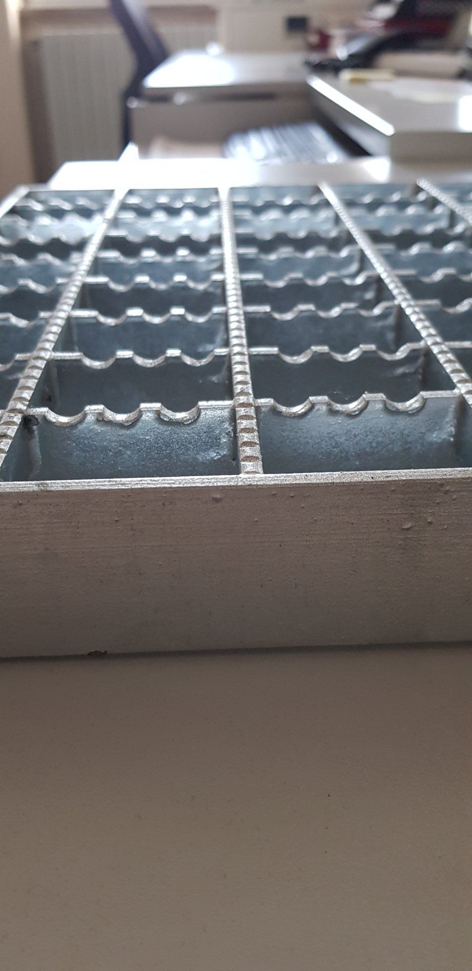 Primo piano di un pannello a griglia metallica su una mensola bianca, con una stanza sfocata sullo sfondo.