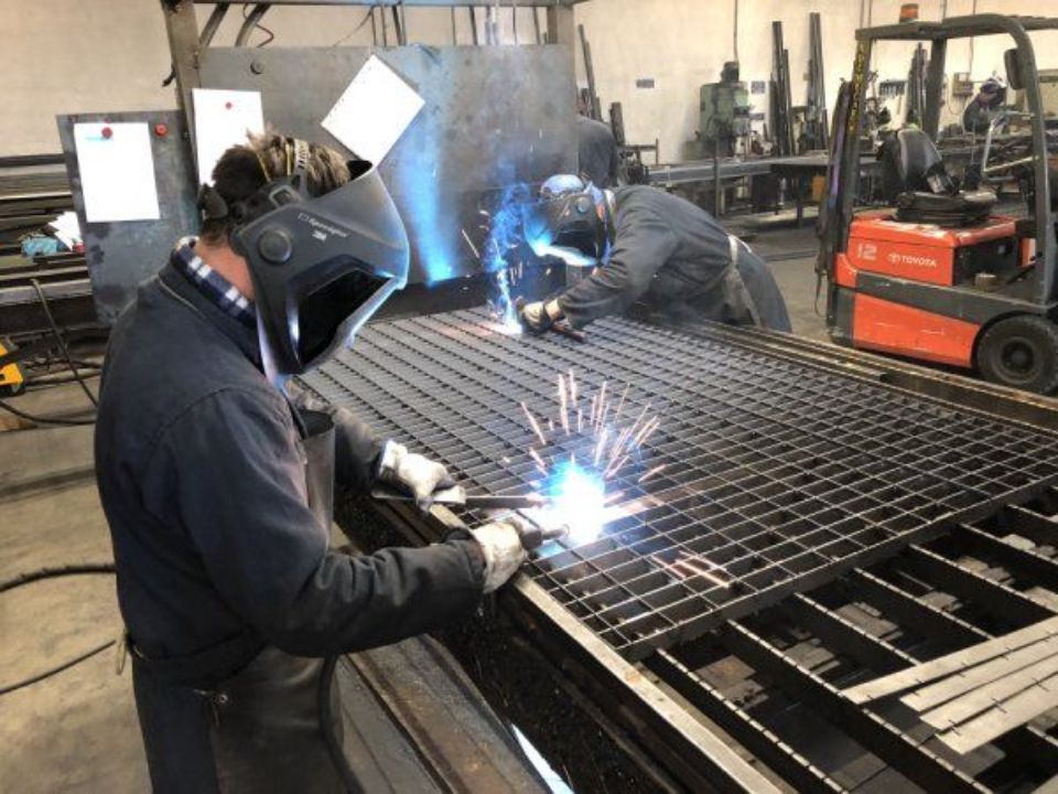 Due operai saldano una griglia metallica in un'officina, tra scintille luminose e con indosso maschere protettive.