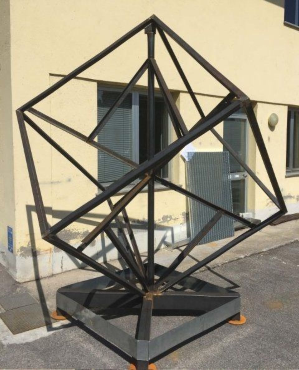Scultura geometrica in metallo nero su una base, posta di fronte alla parete beige di un edificio.