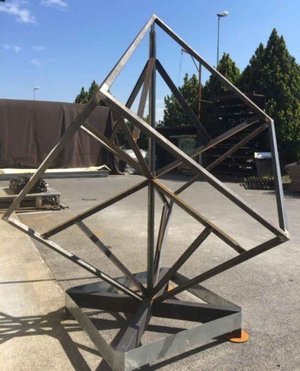 Scultura geometrica in metallo installata all'aperto sul marciapiede, realizzata con barre d'argento che formano una struttura a forma di diamante.