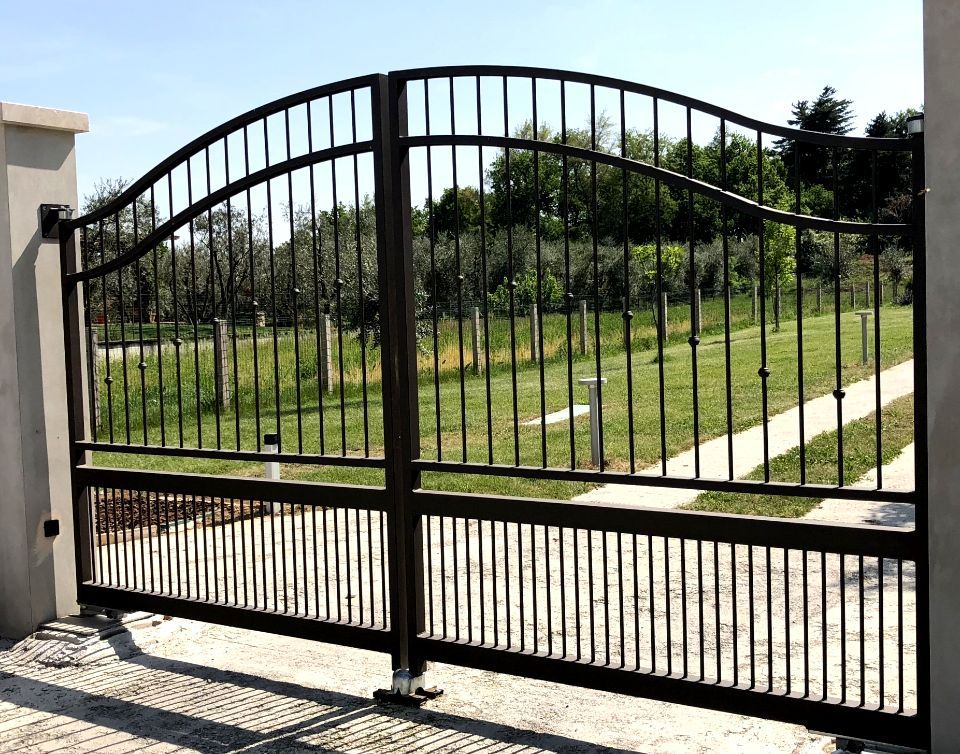 Cancello in metallo nero che si apre su un vialetto erboso e soleggiato e su un cortile.