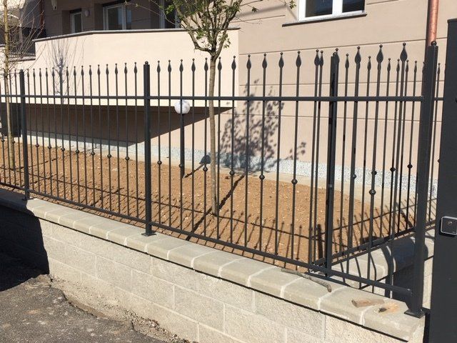 Recinzione in metallo nero lungo un edificio beige accanto a un vialetto pavimentato e a un muro di contenimento.