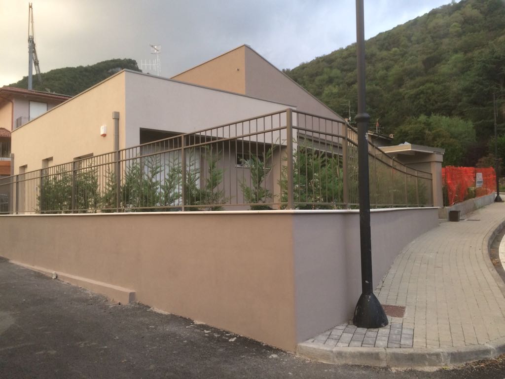 Edificio moderno di colore beige con patio recintato adiacente a un marciapiede lastricato e a una strada in salita.