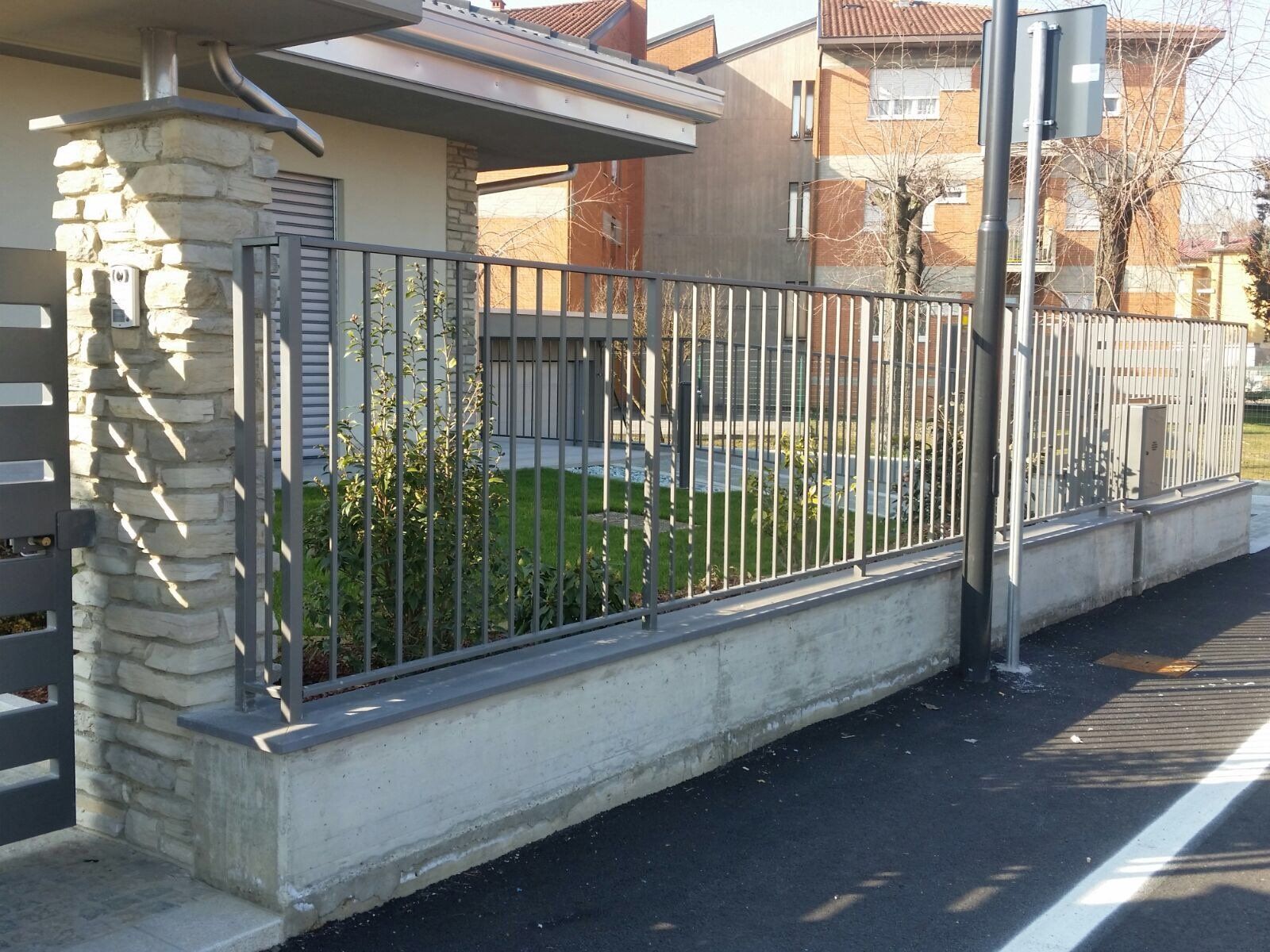 Casa residenziale dietro una recinzione metallica e un basso muro di pietra lungo una strada, alla luce del giorno