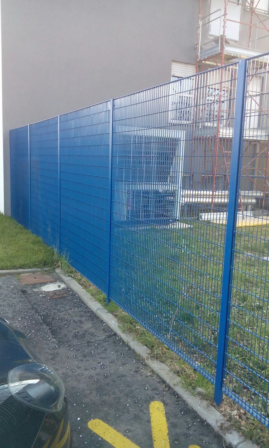 Una recinzione blu a rete metallica costeggia uno stretto passaggio pedonale accanto a un edificio e a un'auto parcheggiata.