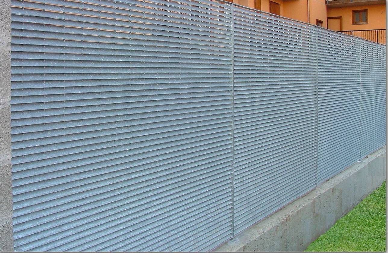 Recinzione in rete metallica blu lungo una base di cemento accanto a un prato e a un edificio arancione.
