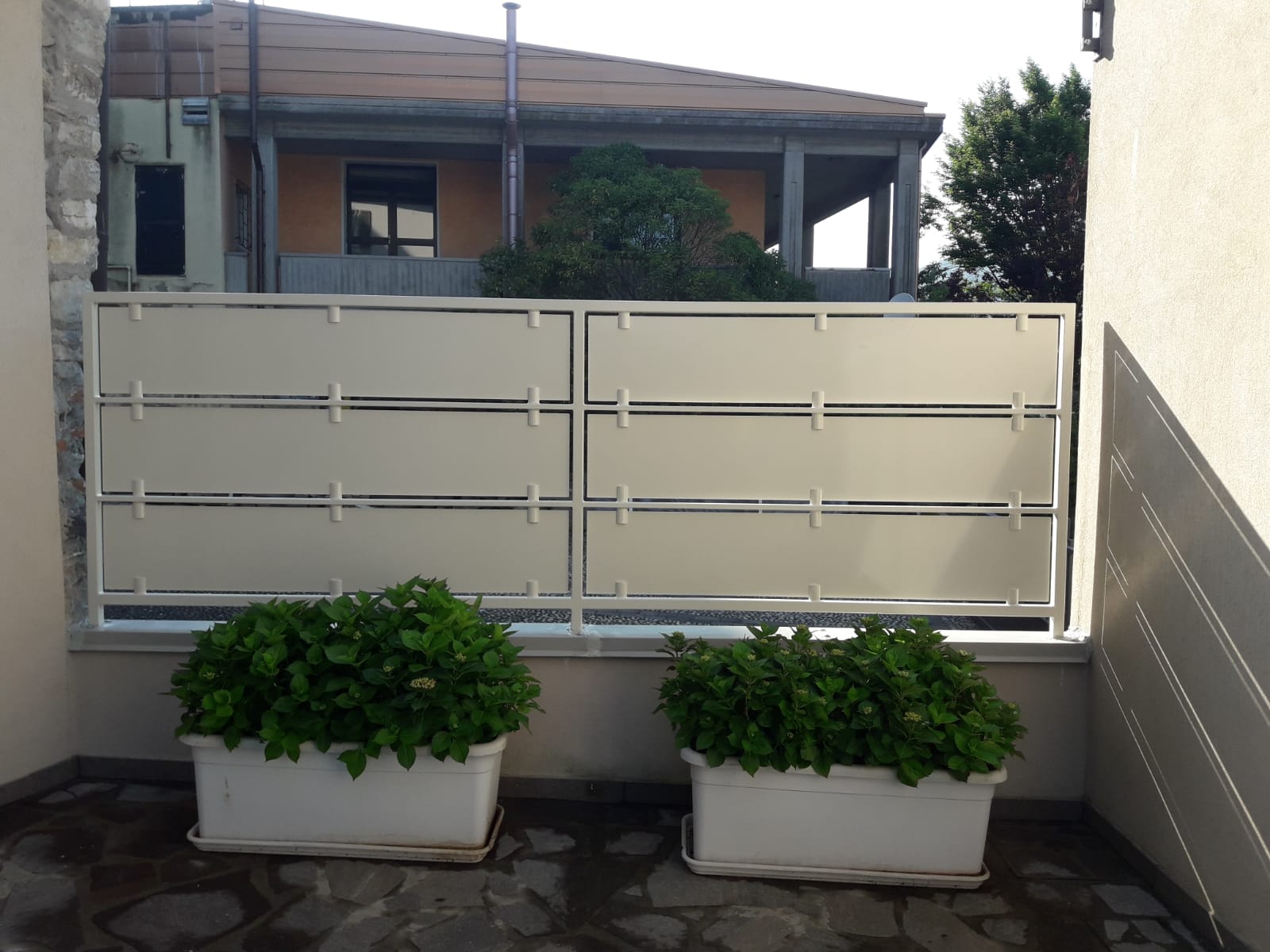 Balcone con due fioriere bianche con arbusti verdi e un paravento beige che si affaccia sulle case vicine.