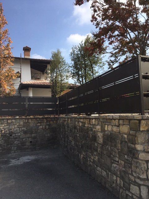 Muro di contenimento in pietra e recinzione nera accanto a una casa con alberi autunnali e cielo azzurro.