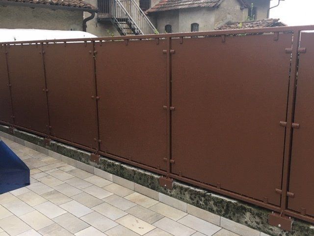 Recinzione metallica marrone lungo un vialetto piastrellato accanto al cortile di un edificio.
