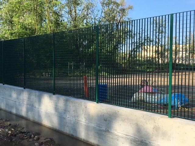 Una recinzione metallica verde accanto a un muro di cemento circonda un piccolo parco giochi con attrezzature colorate e alberi sullo sfondo.