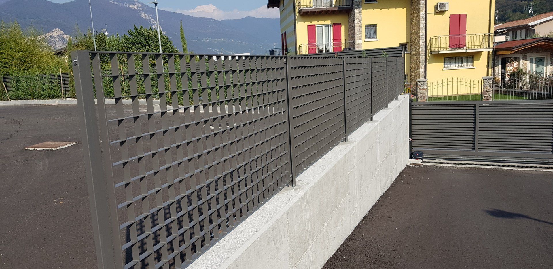 Recinzione metallica scura con base in cemento bianco a lato di una strada, con case gialle e montagne sullo sfondo.