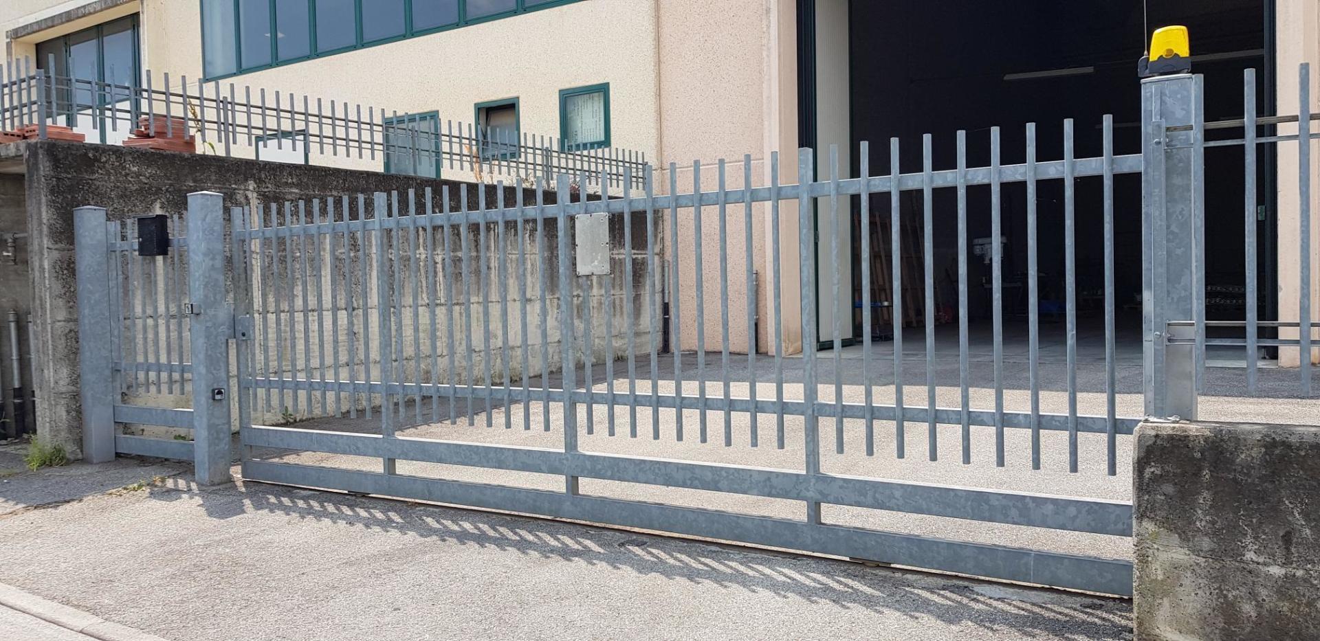 Recinzione e cancello in metallo grigio davanti a un edificio beige con ingresso aperto, su un terreno ghiaioso.