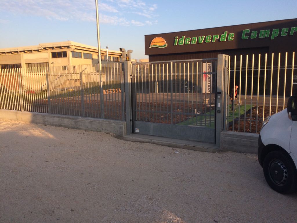 Ingresso di un'attività commerciale con cancello, insegna, recinzione metallica e un furgone bianco parcheggiato al tramonto.