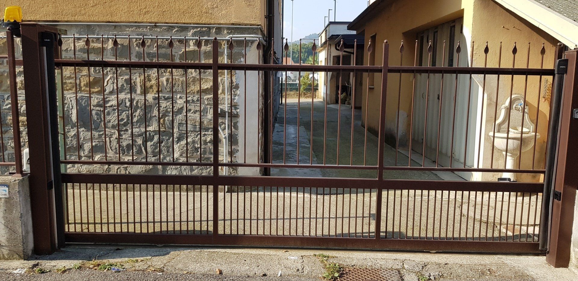 Cancello metallico chiuso tra una strada e uno stretto passaggio pedonale accanto a un edificio giallo.