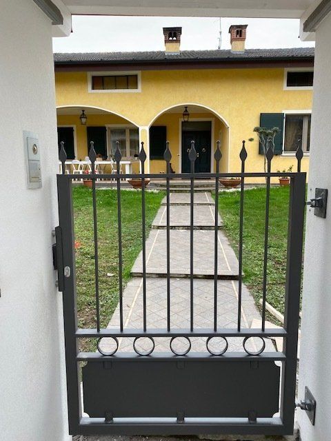 Un cancello di metallo nero si apre su una casa gialla con un vialetto d'ingresso e un prato verde.