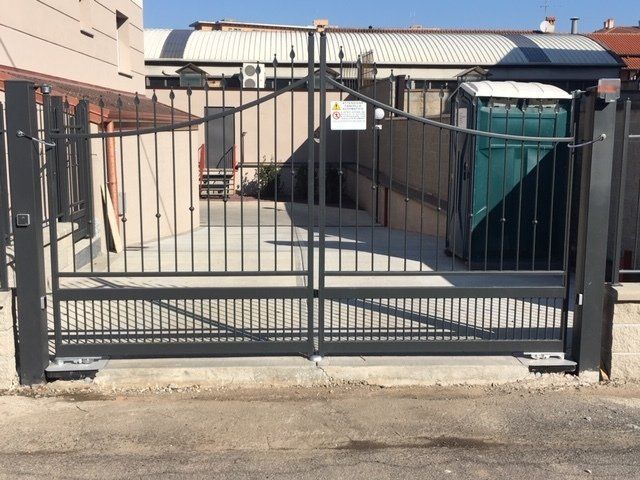 Cancello metallico d'ingresso che blocca uno stretto vicolo accanto a un edificio beige e a un container verde.