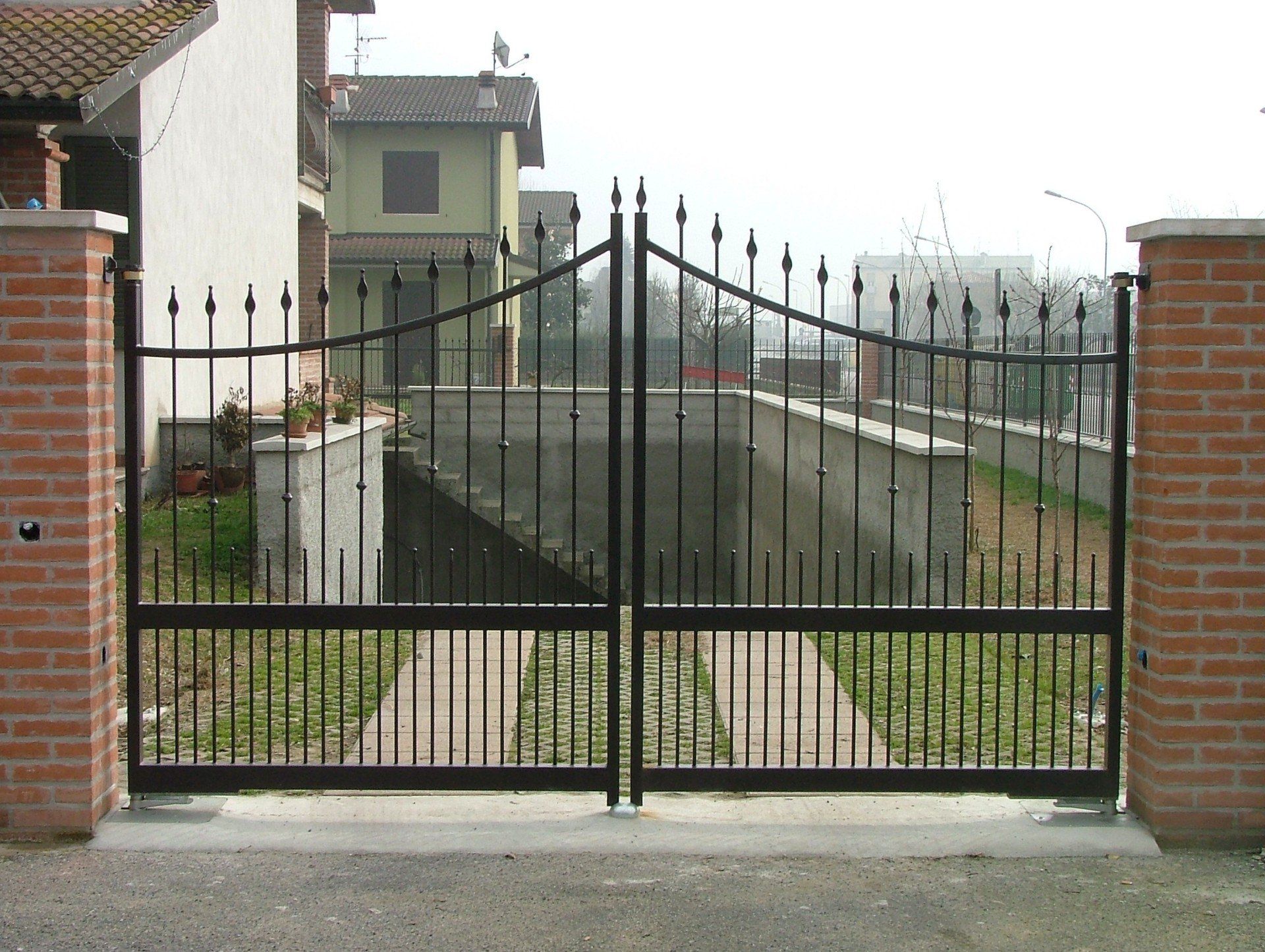Cancello in metallo nero tra pilastri di mattoni di fronte a una casa e al vialetto d'accesso.