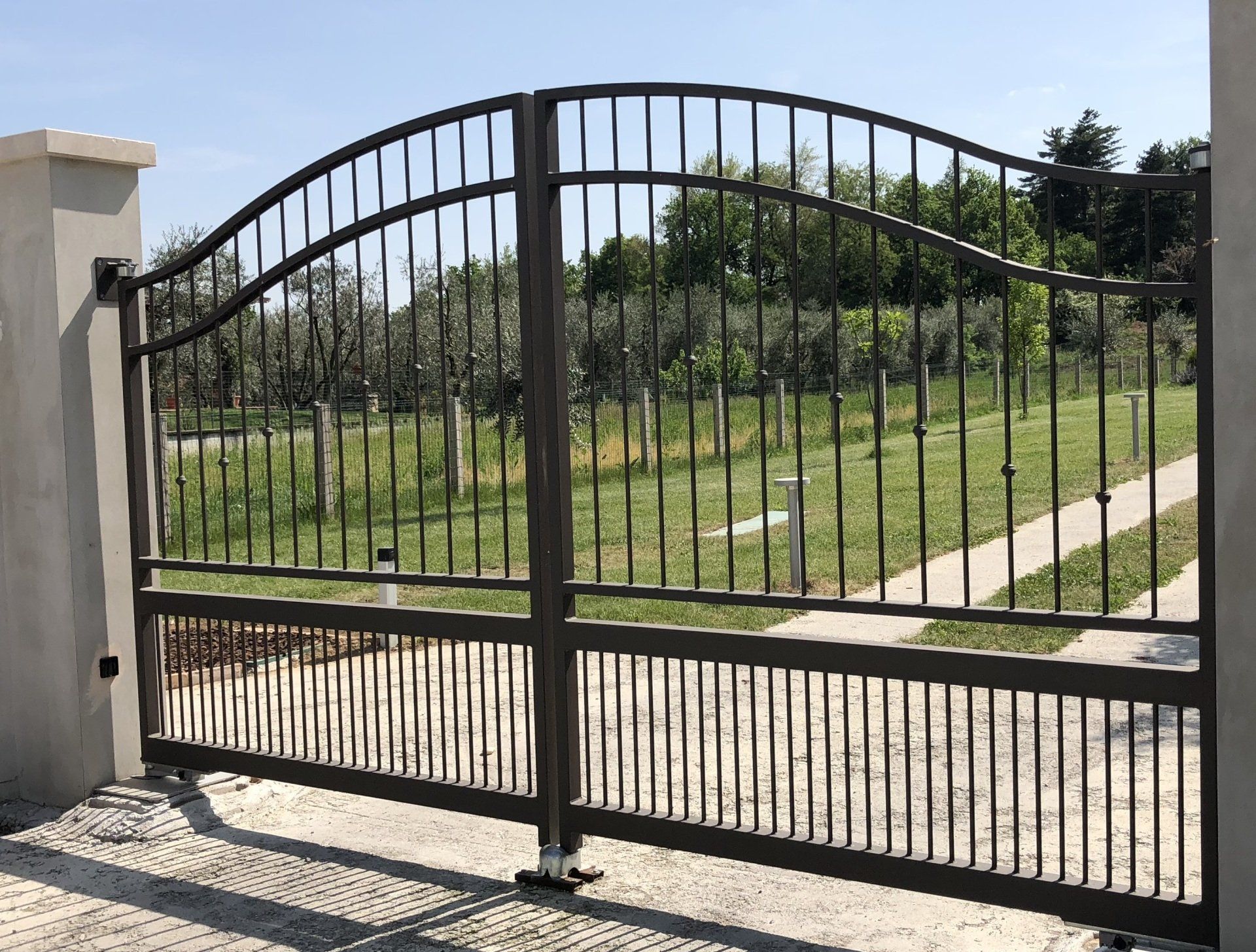Cancello carrabile in metallo nero accanto a un pilastro di cemento, che si apre su un prato e un sentiero.
