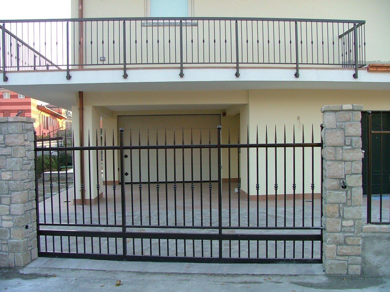 Ingresso della casa con cancello in metallo nero, pilastri in pietra e balcone al secondo piano.