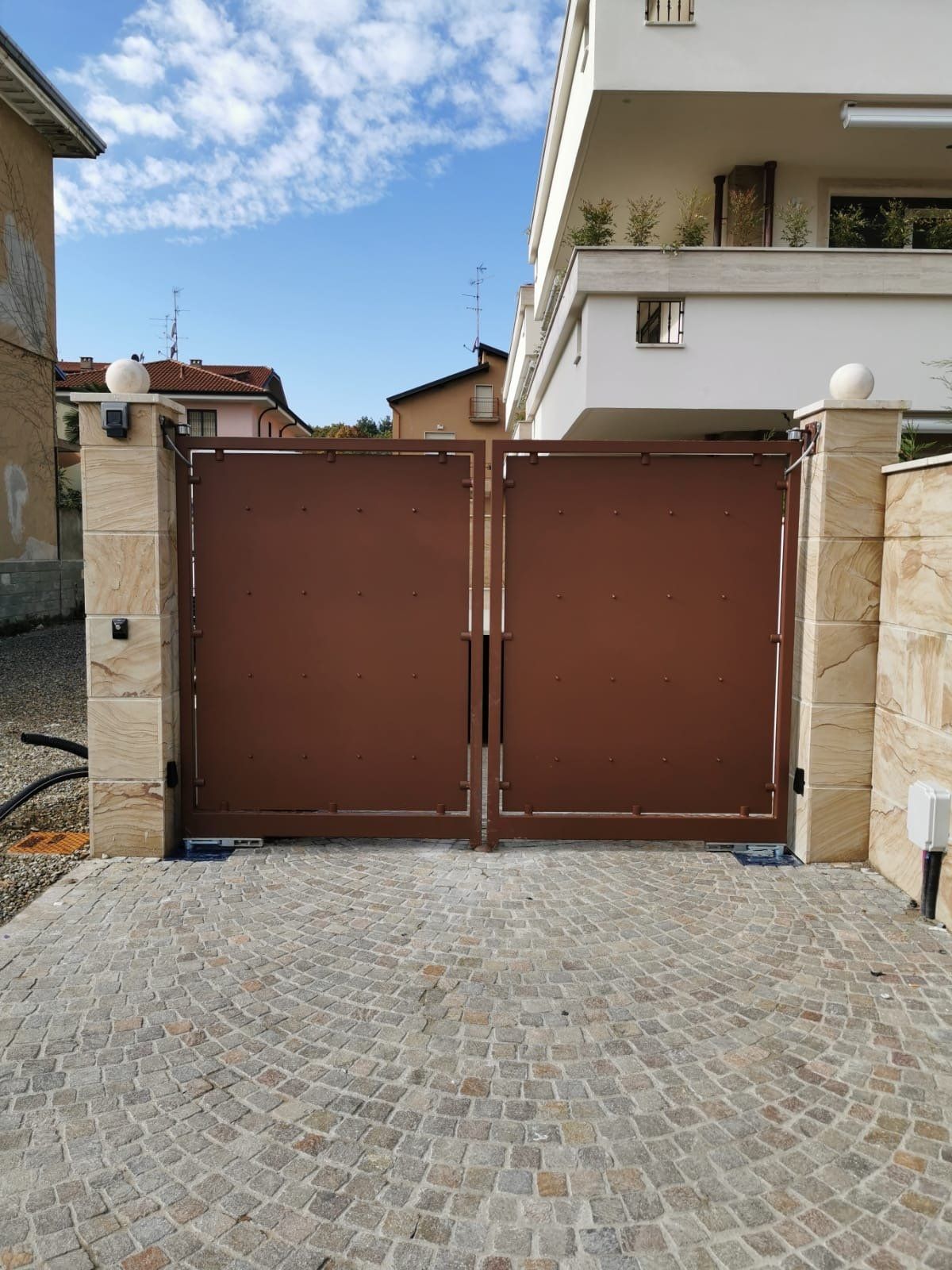 Cancello carrabile in metallo marrone tra pilastri in pietra di fronte a un edificio residenziale.