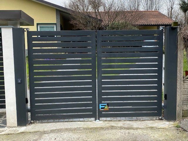 Cancello carrabile in metallo a doghe grigio scuro davanti a una casa con pilastro bianco e tetto di tegole.