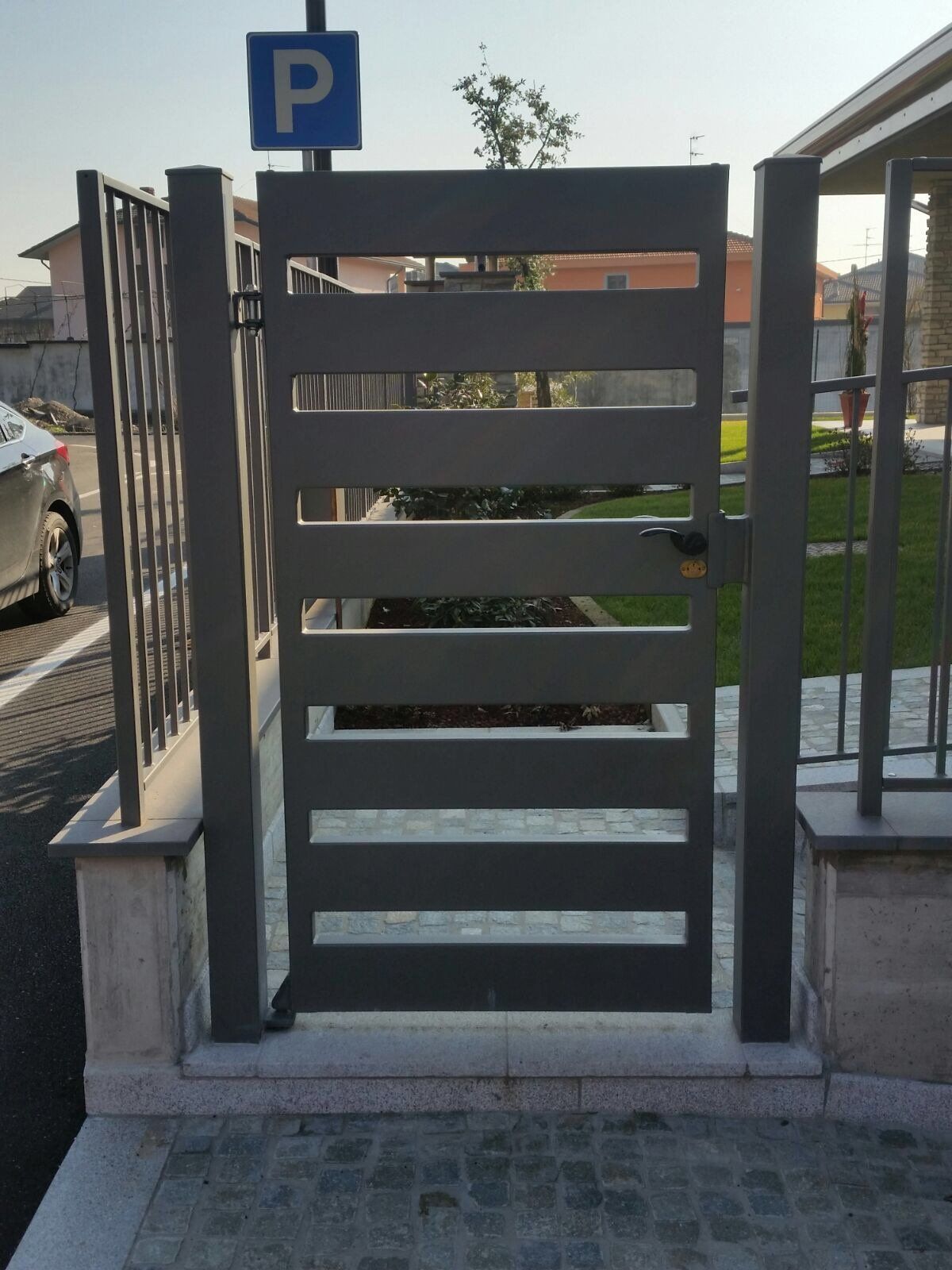 Cancello pedonale verde scuro con doghe orizzontali, situato accanto a un marciapiede e a un cartello di parcheggio.