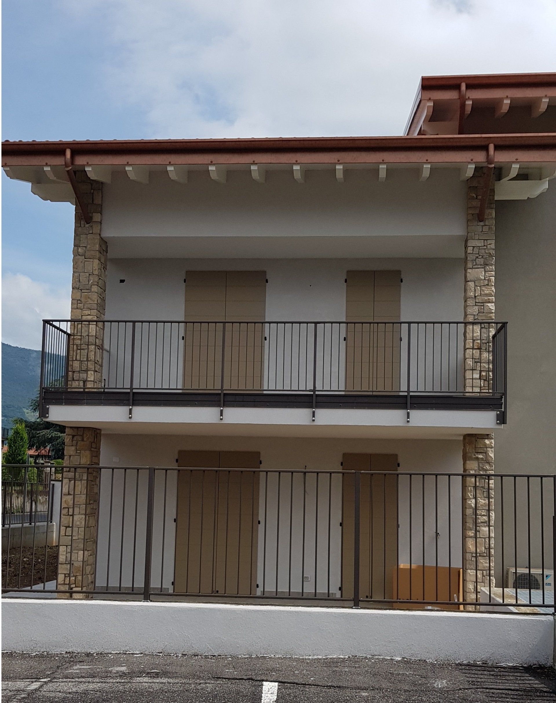 Casa a due piani con balconi, persiane beige, dettagli in pietra e ringhiere in metallo sotto un cielo nuvoloso.