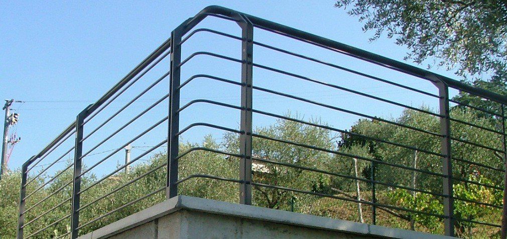 Ringhiera in metallo nero su una sporgenza di cemento, con alberi e cielo azzurro sullo sfondo.