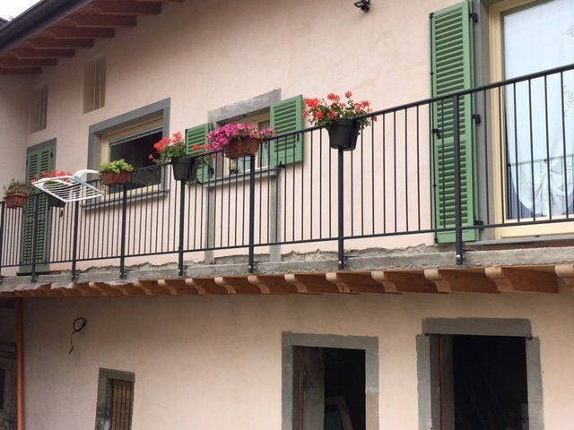 Balcone di una casa a due piani con persiane verdi, fioriere e ringhiere nere.
